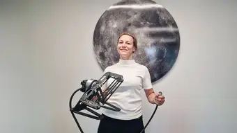 Zdjęcie. Kobieta w średnim wieku o blond włosach ubrana w białą bluzkę stoi na tle fotografii Księżyca trzymając w rękach urządzenie badawcze