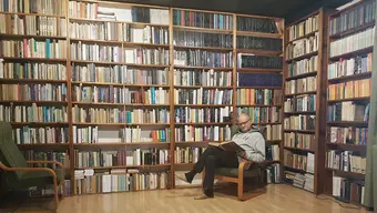 Profesor siedzi w fotelu w bibliotece i czyta książkę na tle regałów wypełnionych książkami.
