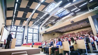 Zdjęcie dużej sali audytoryjnej w któej odbywa się wykład. Po lewej stornie widać prelegenta stojącego na katedrze. Po prawej stronie w rzędach siedza studenci. 
