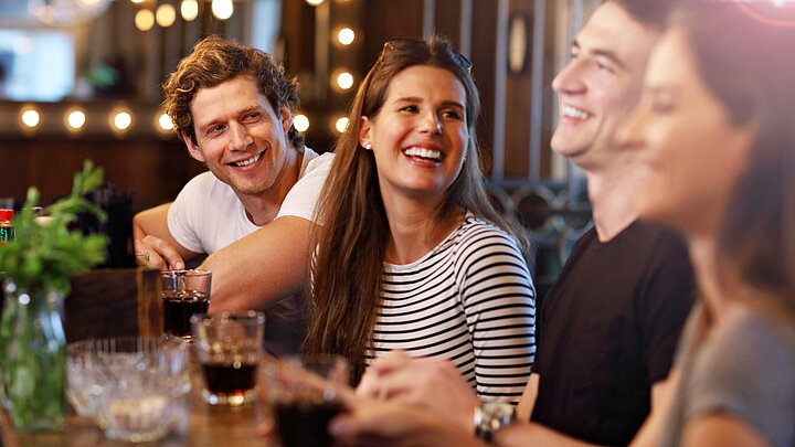 Grafika dekoracyjna. Cztery uśmiechnięte osoby siedzą obok siebie przy długim stole w restauracji. Przed sobą mają szklanki z napojami, śmieją się i rozmawiają