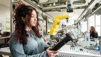 Zdjęcie dekoracyjne. Młoda kobieta w pracowni inżynierskiej; trzyma w dłoniach przedmiot, który wyglądem przypomina panel sterowniczy. Za kobietą duży stół inżyniersko-projektowy z rozstawionym oprzyrządowaniem elektroniczno-technicznym (m.in. sporych rozmiarów chwytak w kolorze żółtym, podpięte kable). Za stołem siedzi jedna osoba. Nad stołem suwnica.